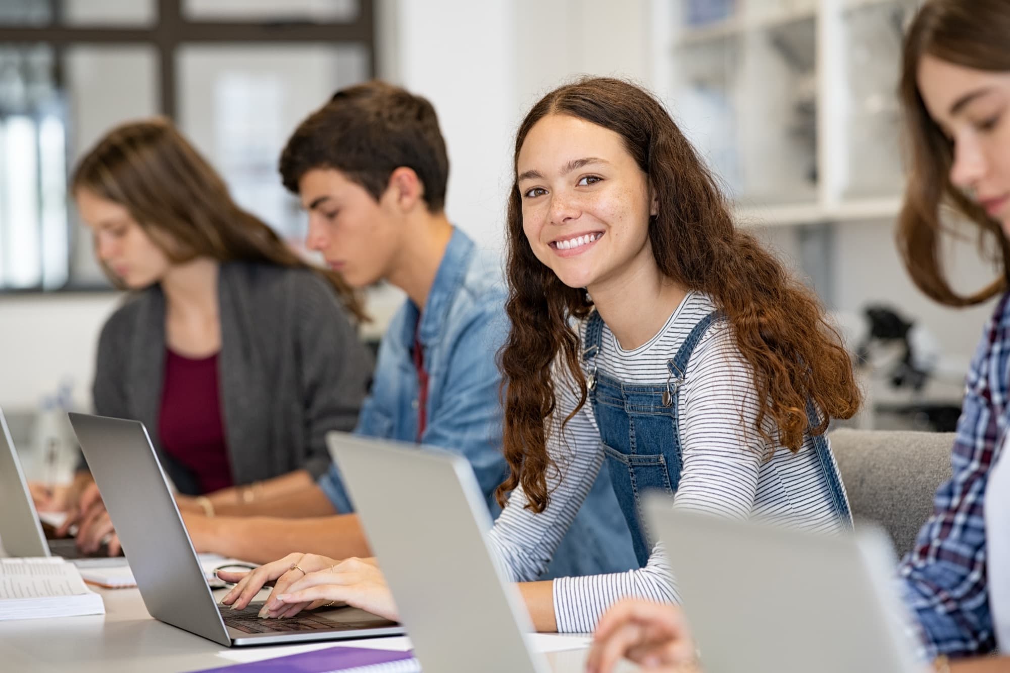 Smiling student with long curly hair at a row of laptops in a bright classroom