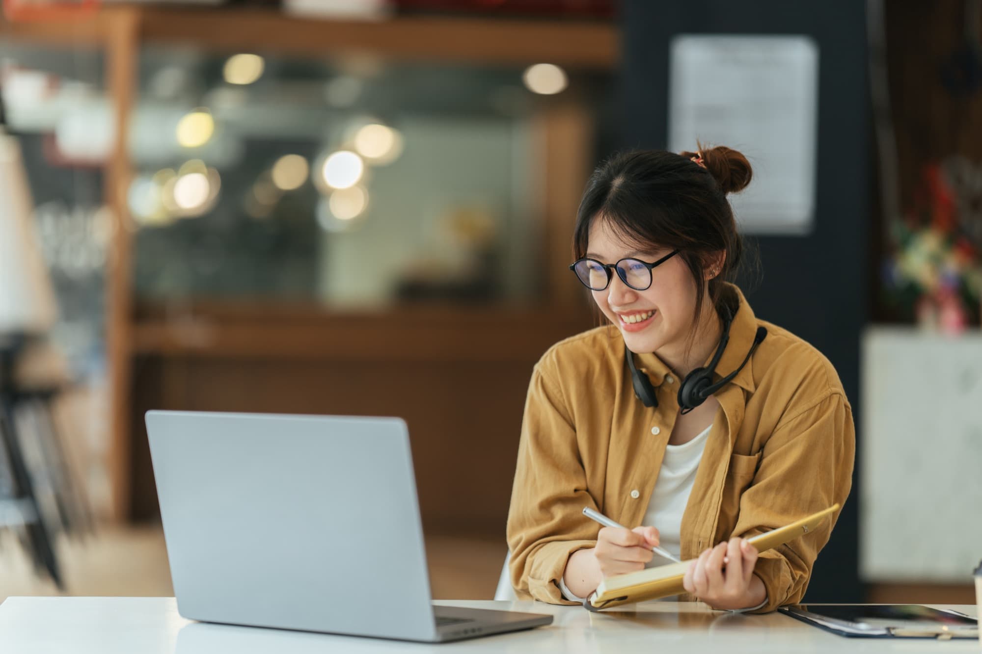 Smiling student in glasses with a headset around her neck, writing in a notebook beside a laptop