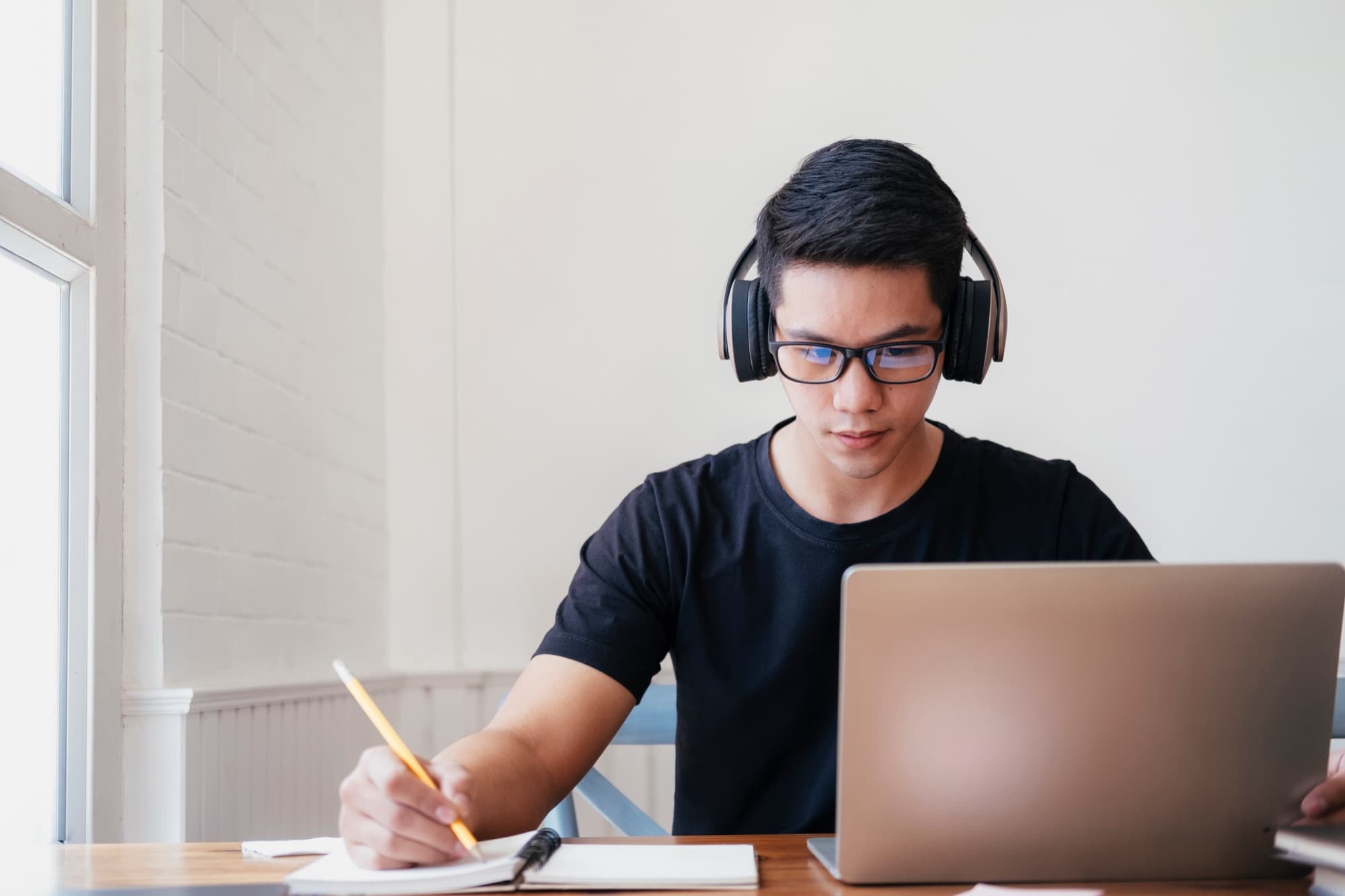 Student wearing headphones takes notes with a pencil beside his laptop